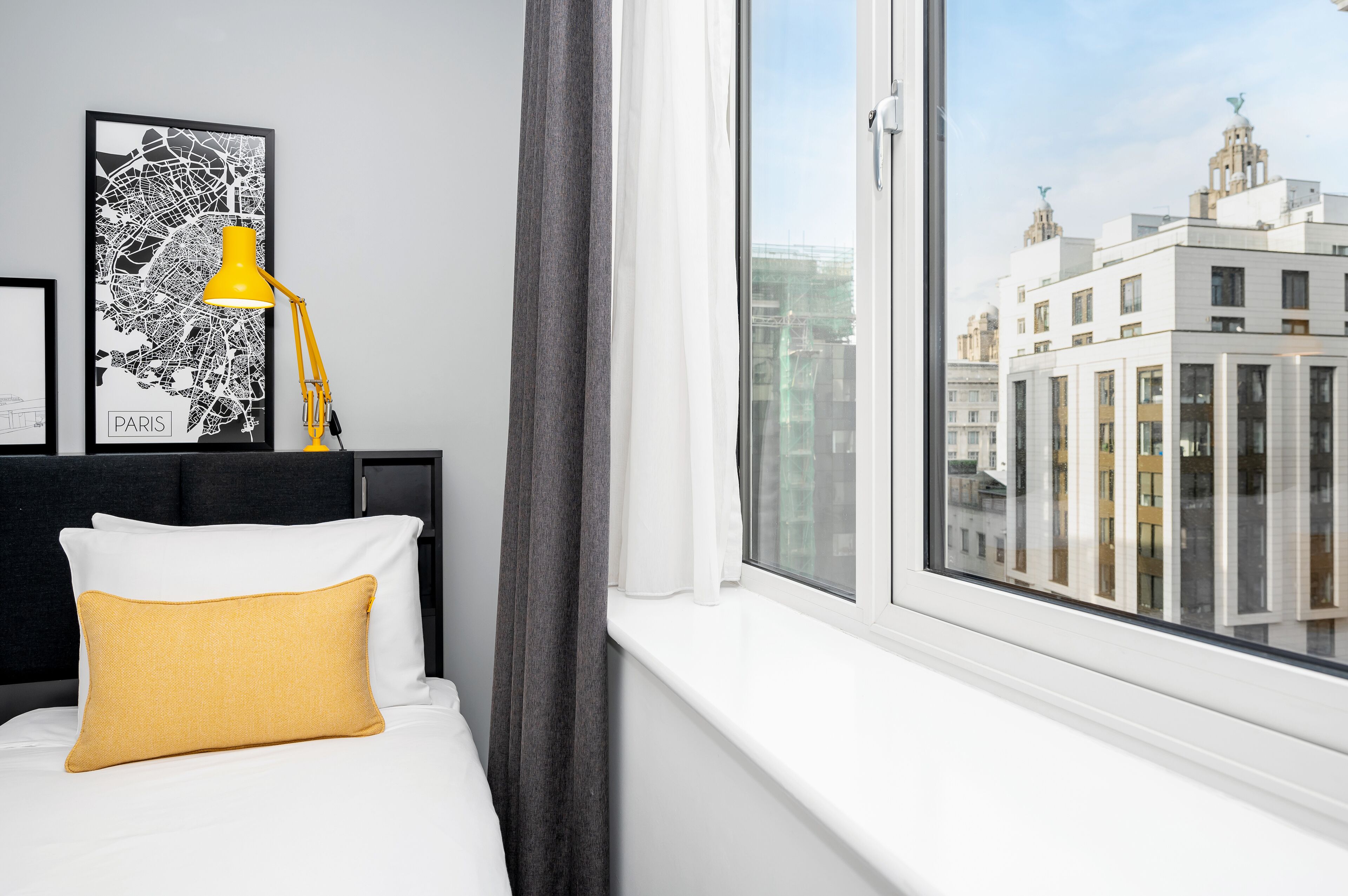 Ett hotellrum med en säng, en gul lampa, en svartvit karta över Paris och utsikt över stadens byggnader.