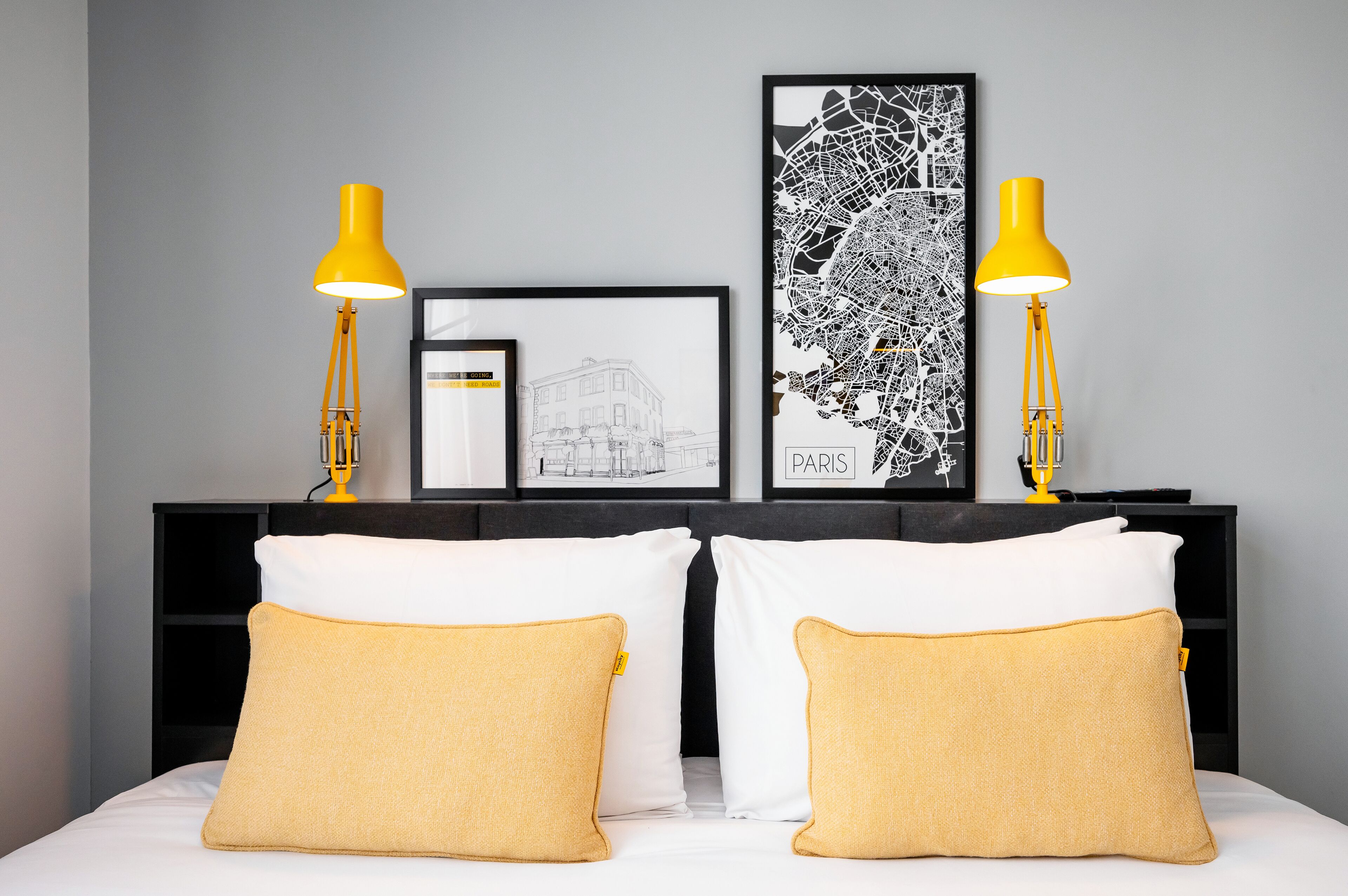 Una cama bien hecha con almohadas blancas y amarillas, dos lámparas de noche amarillas y obras de arte en un cabecero oscuro.