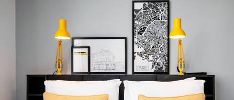 A neatly made bed with white and yellow pillows, two yellow bedside lamps, and framed artwork on a dark headboard.