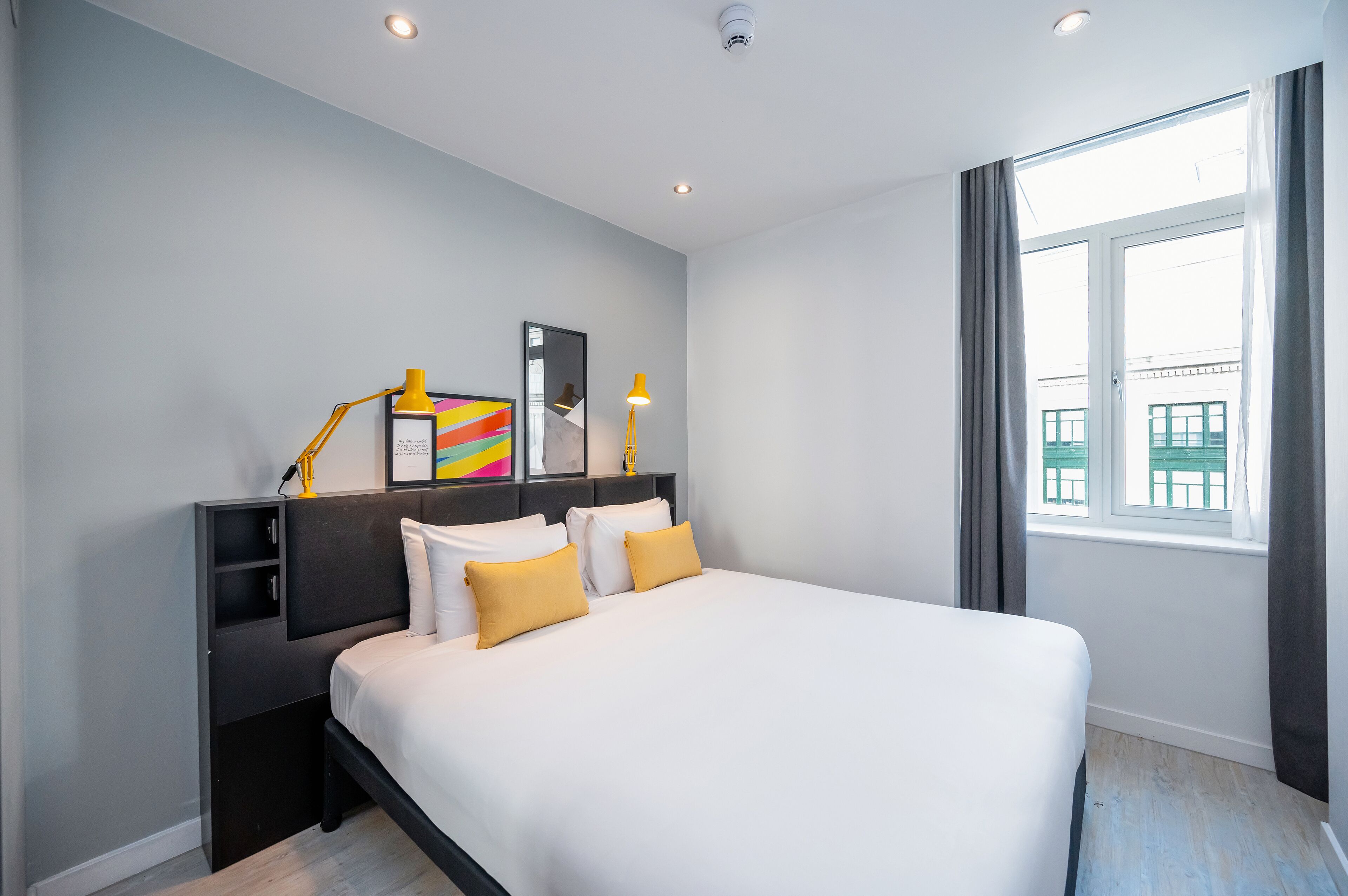 A modern hotel room with a large bed, bedside tables, a mirror, and a window with curtains.