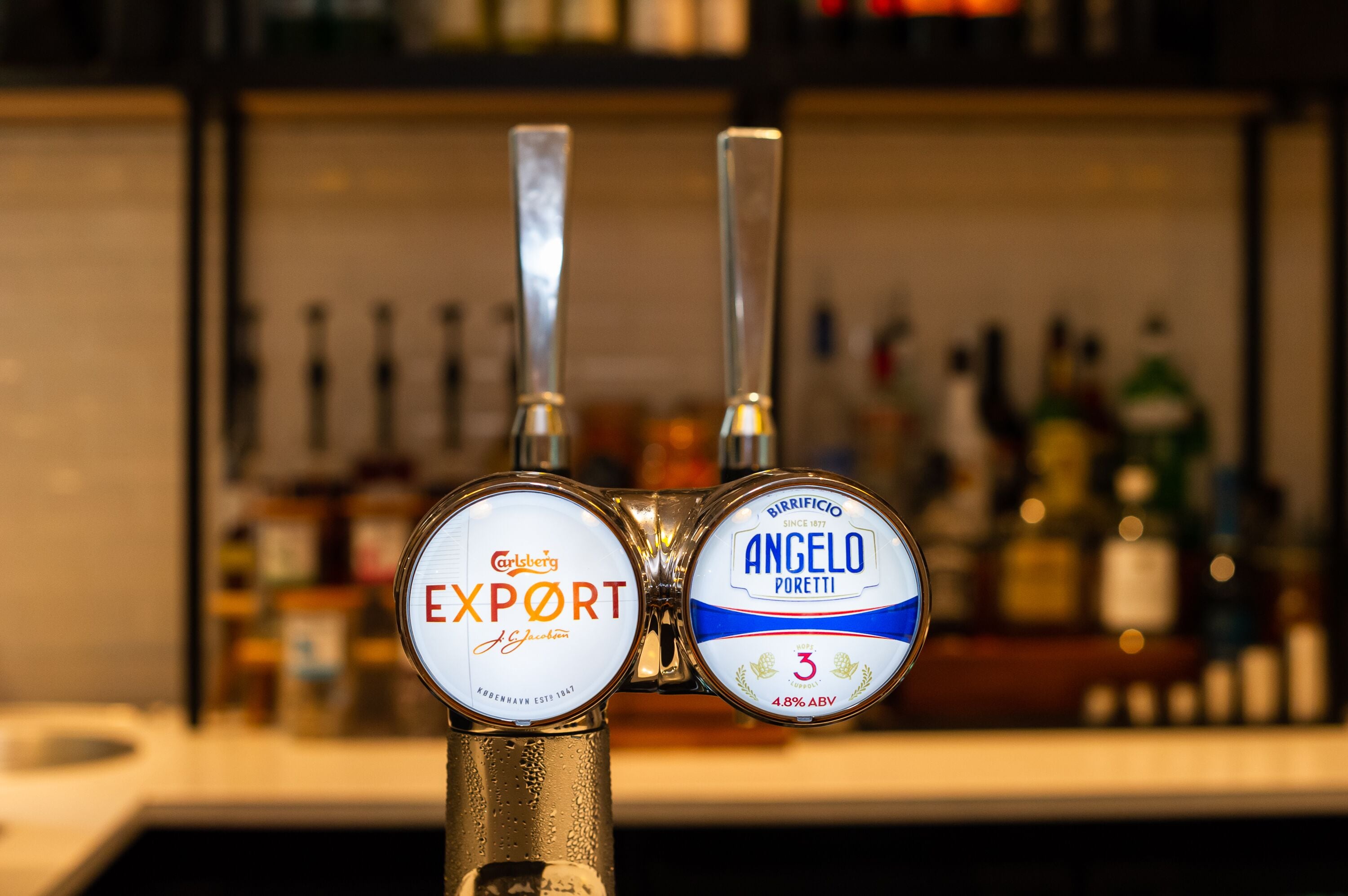 A tap with two beer labels, one for Carlsberg Export and one for Angelo Poretti, with a blurred background of bottles.