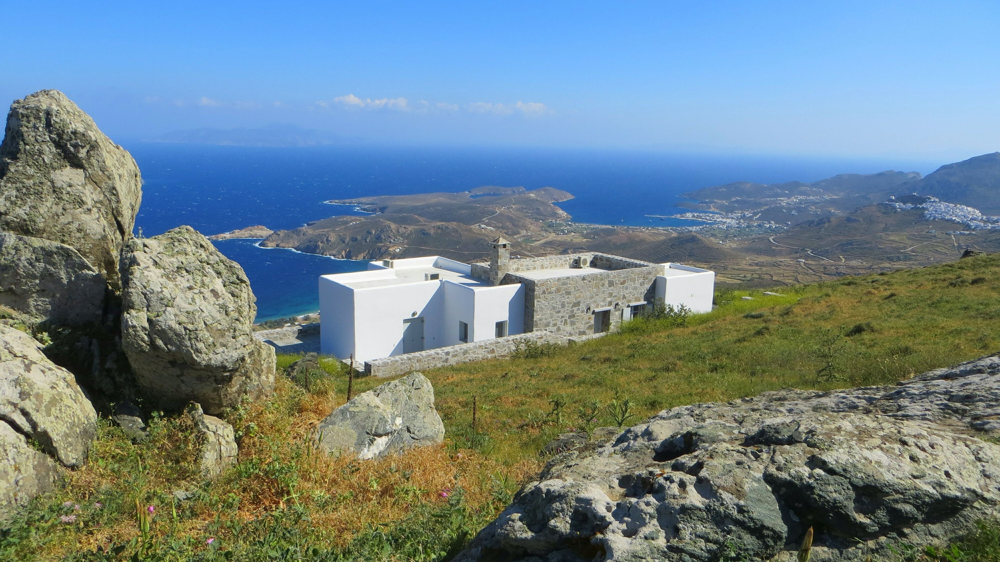 Um edifício branco de paredes de pedra, rodeado por terreno rochoso e com vista para a paisagem costeira.
