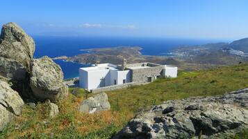Um edifício branco de paredes de pedra, rodeado por terreno rochoso e com vista para a paisagem costeira.