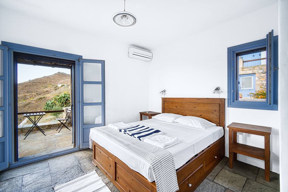 Ein Schlafzimmer mit einem Holzbett, einem kleinen Nachttisch, einem Fenster mit Blick auf eine Hügelandschaft und einer offenen Tür, die auf eine Terrasse mit Tisch und Stühlen führt.