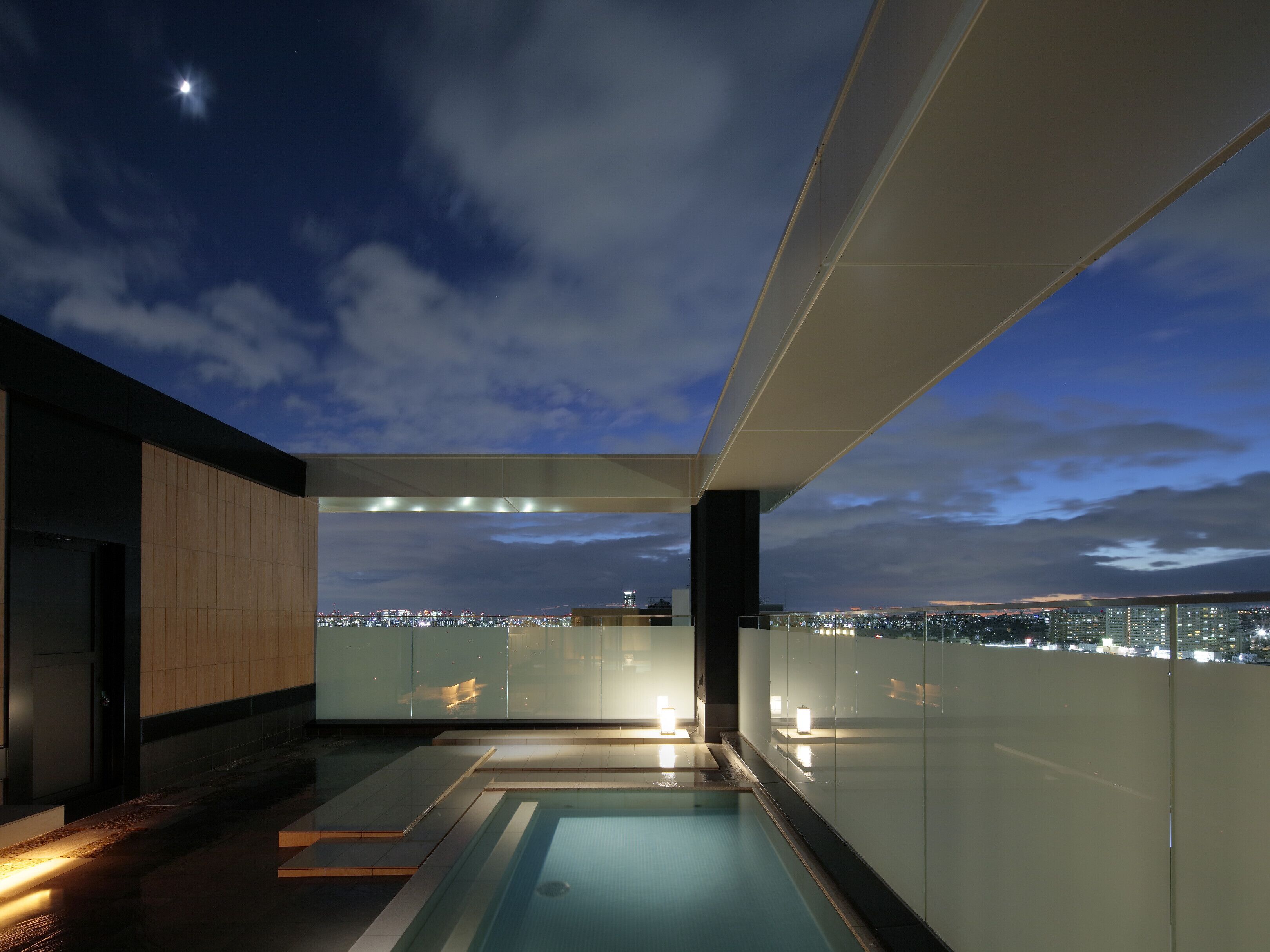 A modern rooftop pool with glass railings, a cityscape view, and a clear night sky.