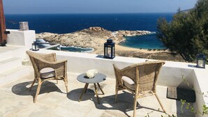 Eine Terrasse mit Korbstühlen und einem Tisch mit Blick auf eine Küstenlandschaft mit felsigem Gelände und dem Meer.