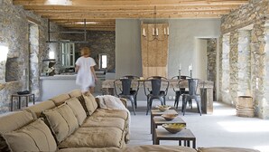 Ein rustikales Interieur mit einer Steinwand, Holzbalkendecke, einer Couch, einem Esstisch, Stühlen und einem Kronleuchter.