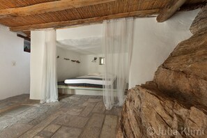 Une chambre à coucher rustique avec un sol en pierre, un lit recouvert de draps blancs et un plafond en bois.