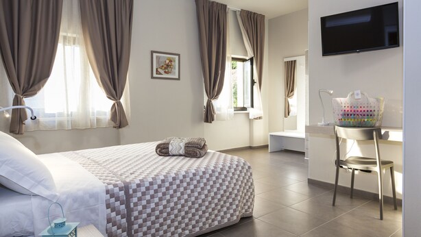 A hotel room with a checkered bedspread, a flat-screen TV mounted on the wall, a desk with a chair, and a window with curtains. - Riviera Hub (Cisano sul Neva)
