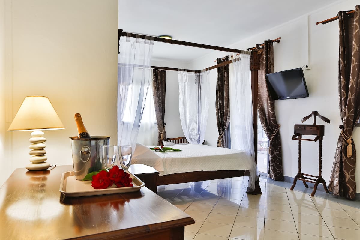 Quarto de hotel com cama, mesa de cabeceira com abajur, bandeja com flores, televisão na parede e um pequeno suporte de madeira.