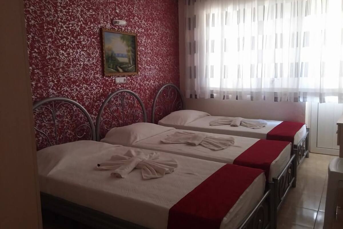 A hotel room with two beds, each with a red bedspread and white towels. There is a painting on the wall and a window with sheer curtains.
