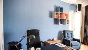 Una habitación con pared azul, una silla de oficina negra, un sillón azul, una mesita con una cesta y una cesta llena de objetos de colores, además de una impresora sobre un escritorio.