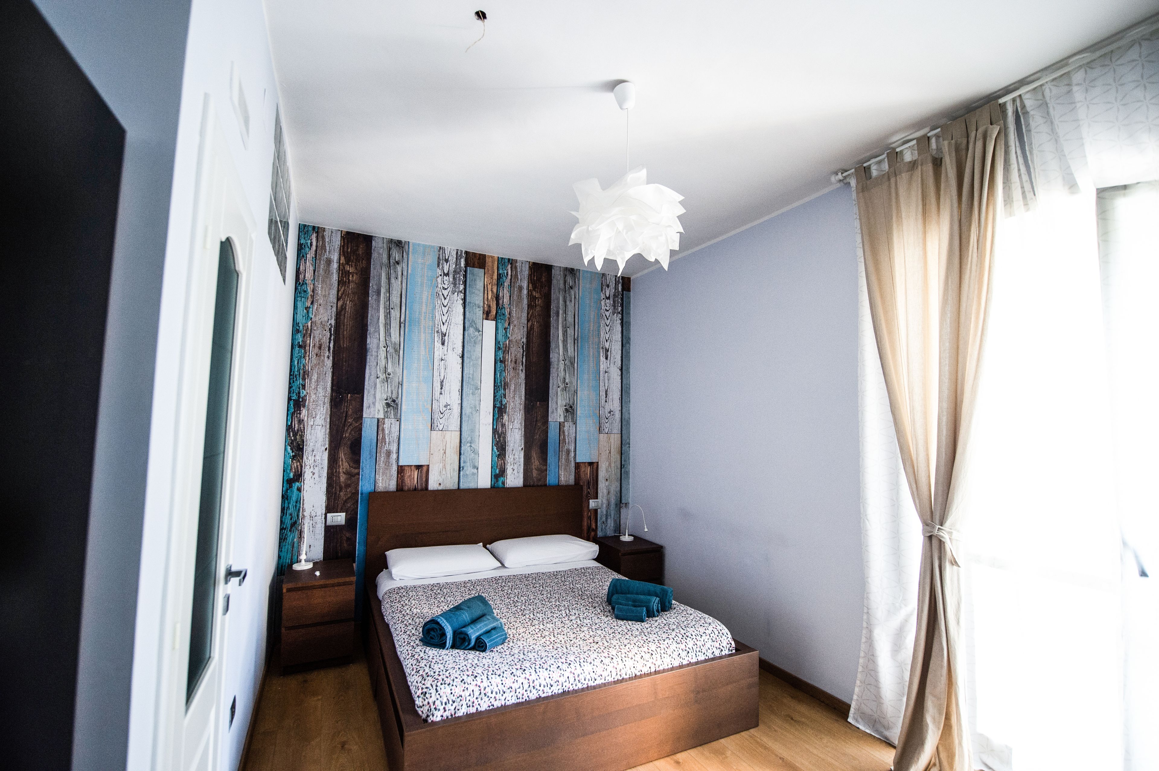 A bedroom with a wooden bed, a white ceiling light, a nightstand, and a wall with a striped pattern.
