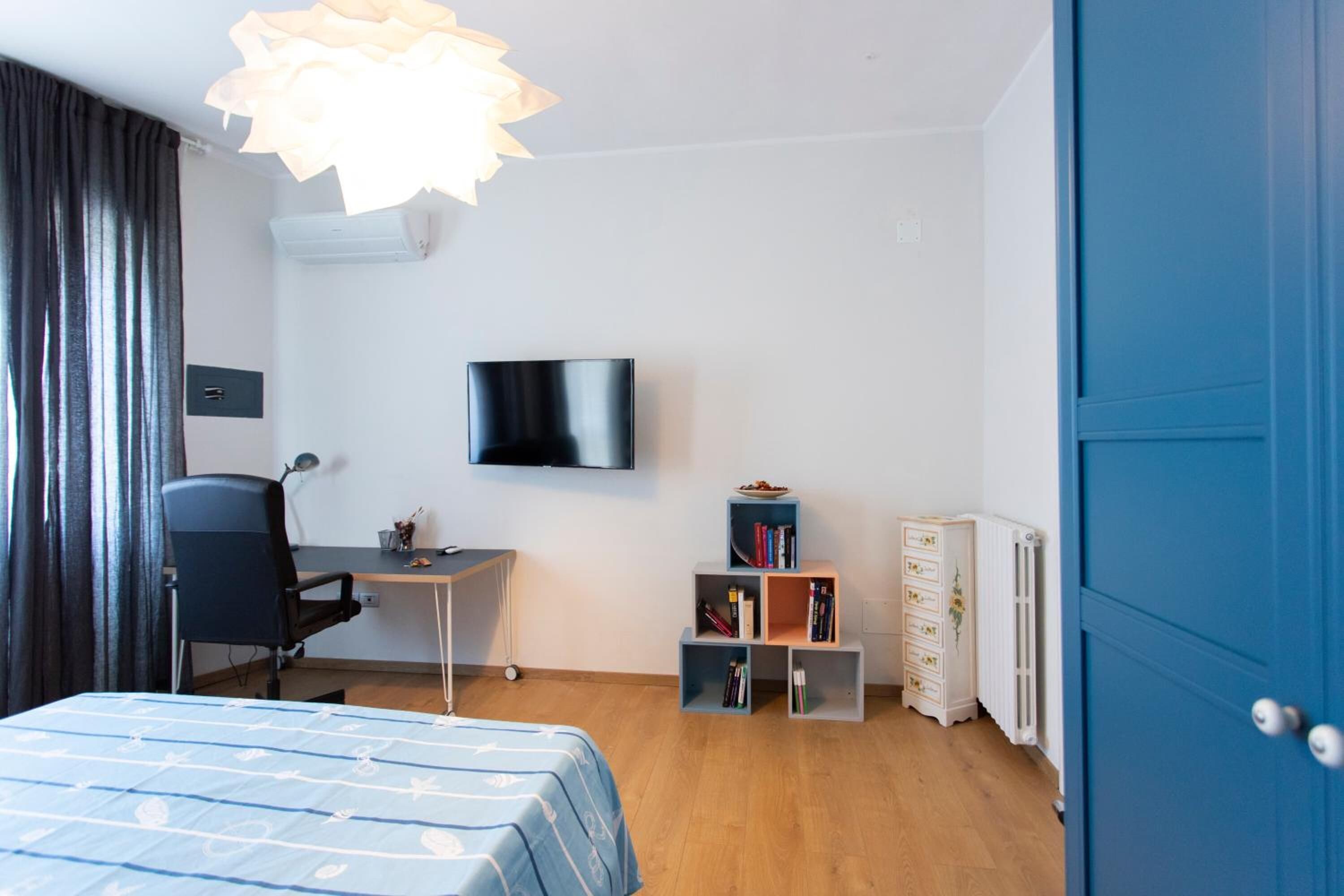 Una habitación con cama, escritorio, silla, televisor, estantería y radiador.