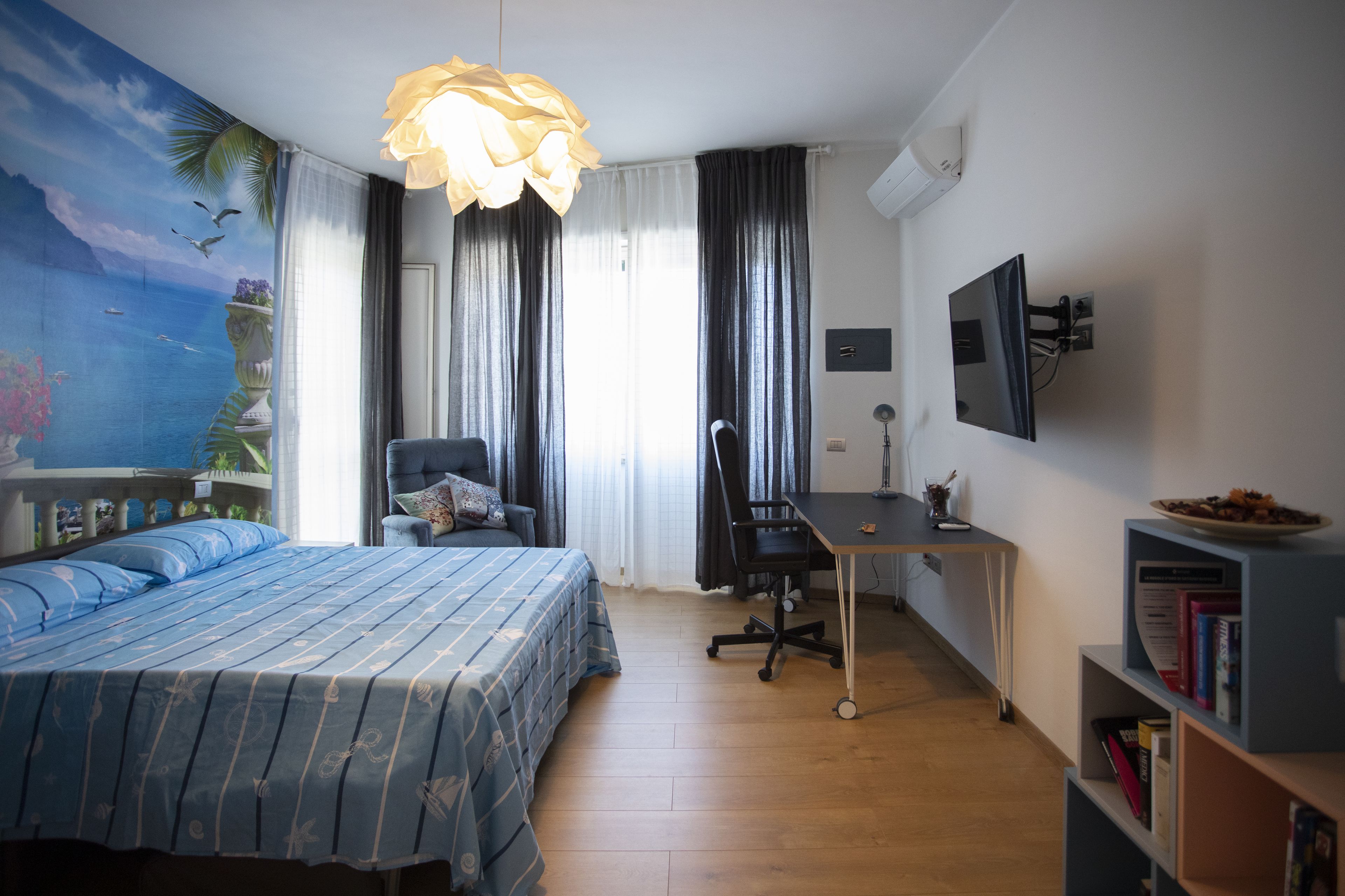 A bedroom with a bed, desk, chair, bookshelf, and a painting of a tropical scene.