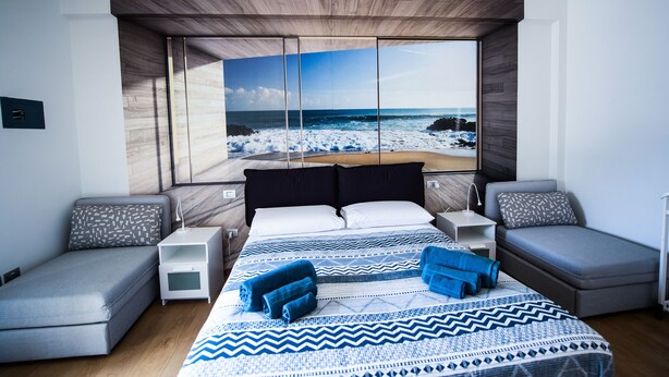 Un dormitorio moderno con una cama grande, dos mesitas de noche, una mesita auxiliar, vistas a la playa y un techo de madera.