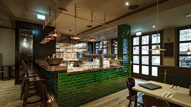 A bar area with a green tiled backsplash, wooden bar stools, and pendant lighting. - Marias Platzl (Munich)