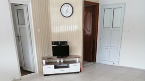 A living room with a wooden coffee table, a yellow sofa, a TV on a wooden stand, and a clock on the wall.