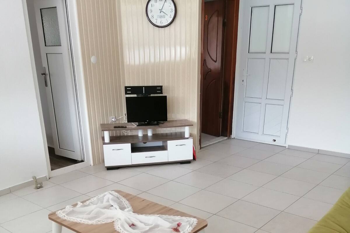 A living room with a wooden coffee table, a yellow sofa, a TV on a wooden stand, and a clock on the wall.
