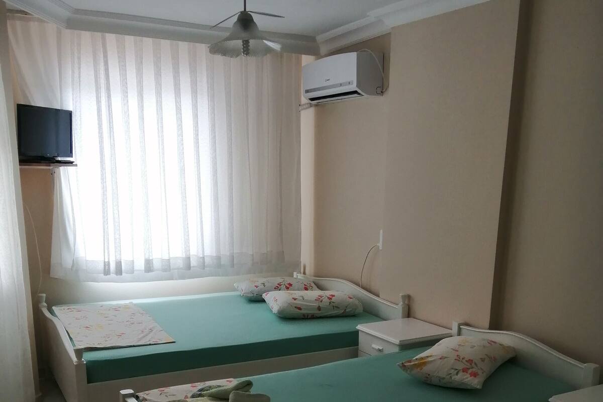 A room with two single beds, a ceiling fan, an air conditioning unit, and a TV mounted on the wall.