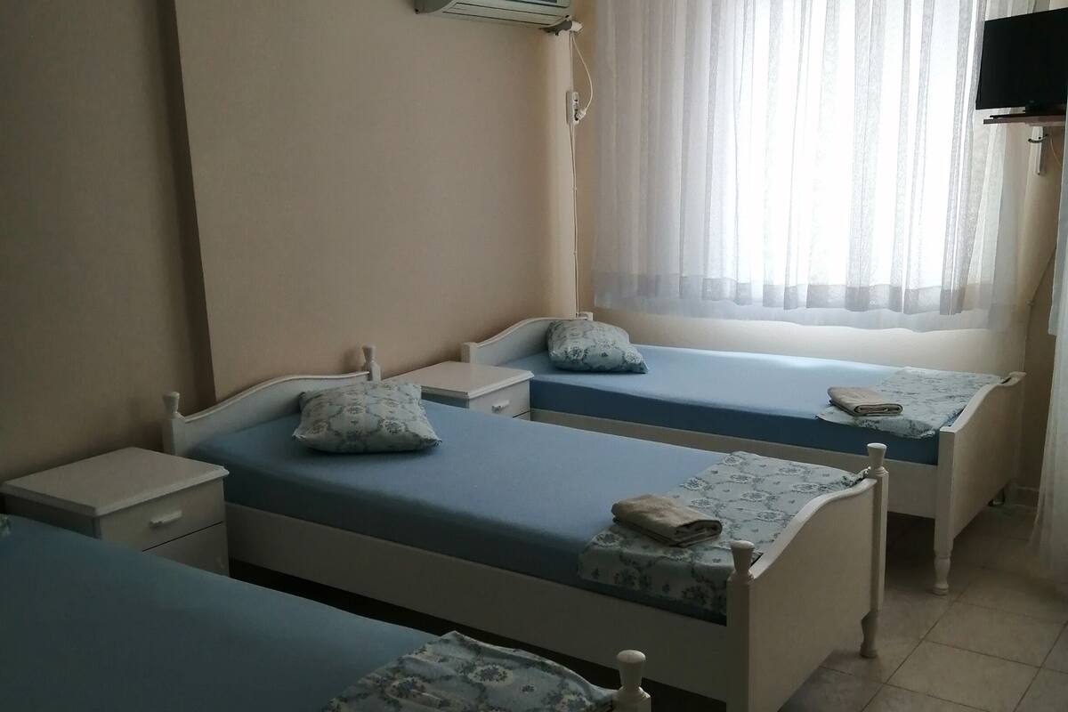 A room with two single beds, a ceiling fan, an air conditioning unit, a window with sheer curtains, and a television mounted on the wall.