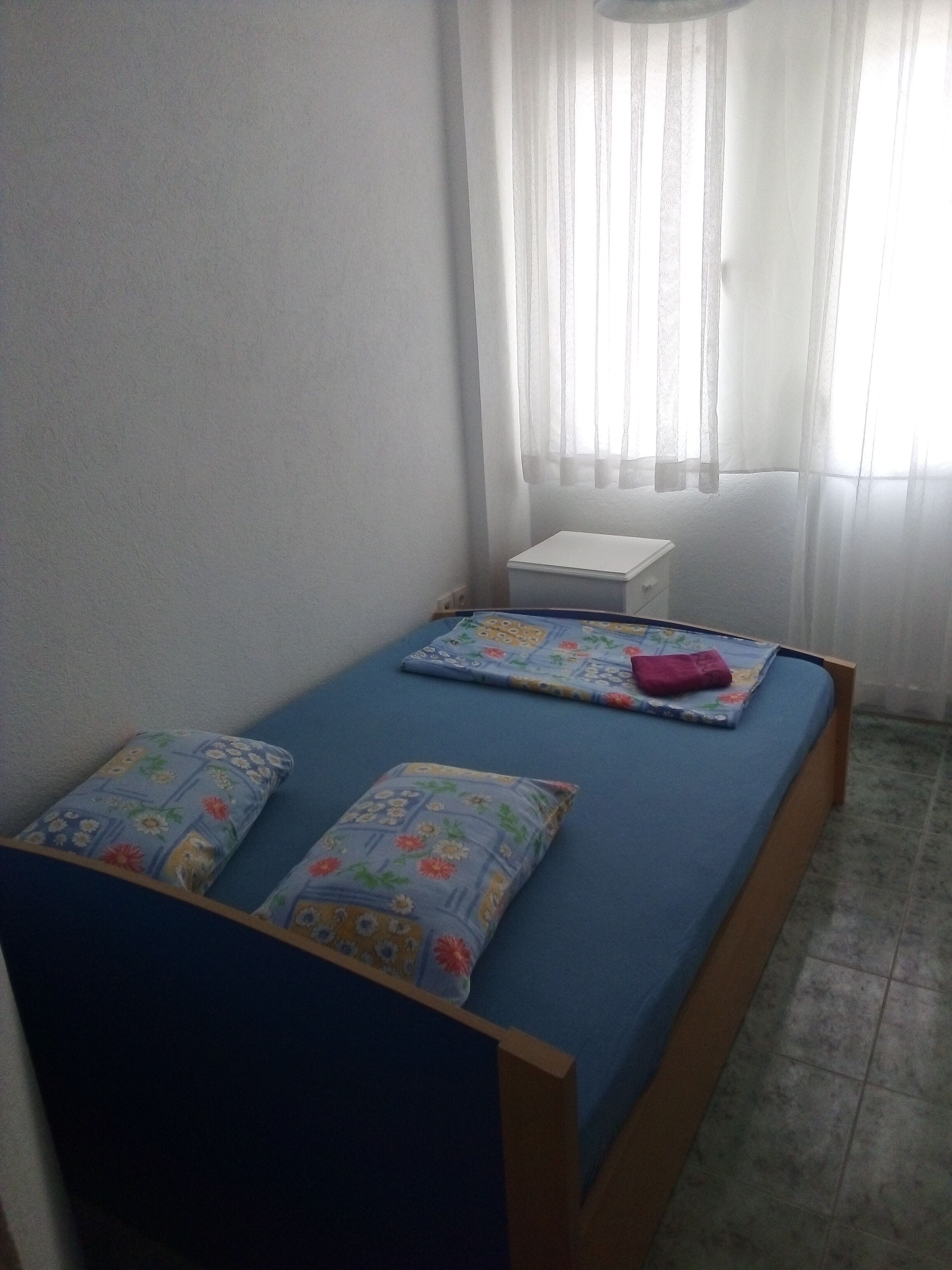 A small, single-bed room with a blue mattress, floral-patterned pillows, a white bedside table, and a window with sheer curtains.