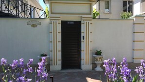 Entrada de um edifício com uma porta preta fechada, uma placa acima e flores roxas em vasos de ambos os lados.