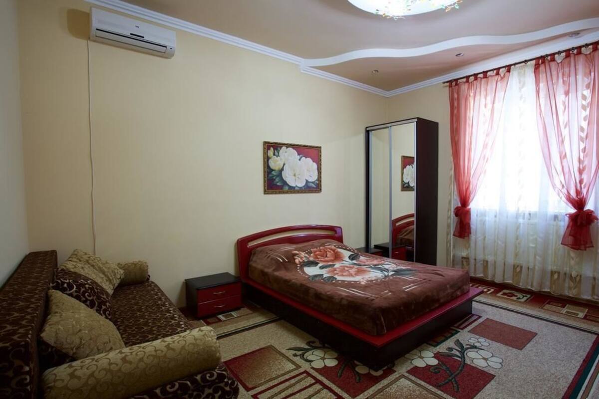 Quarto com cama, sofá, mesa de cabeceira, espelho e janela com cortinas.