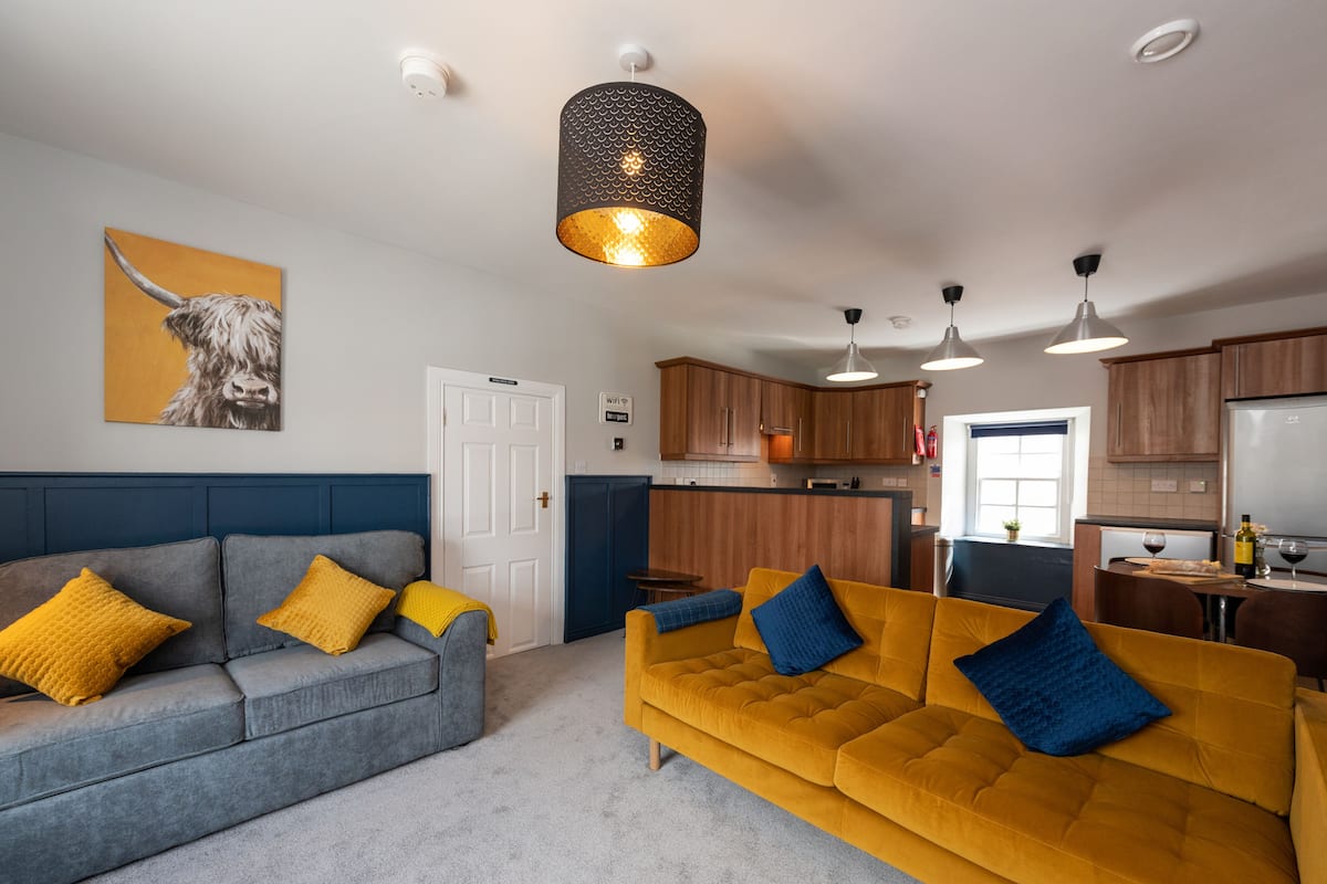 A modern living room with a grey sofa and a mustard sofa, both with yellow and blue cushions. There is a kitchen area with wooden cabinets and a dining table with a wine bottle and glass.
