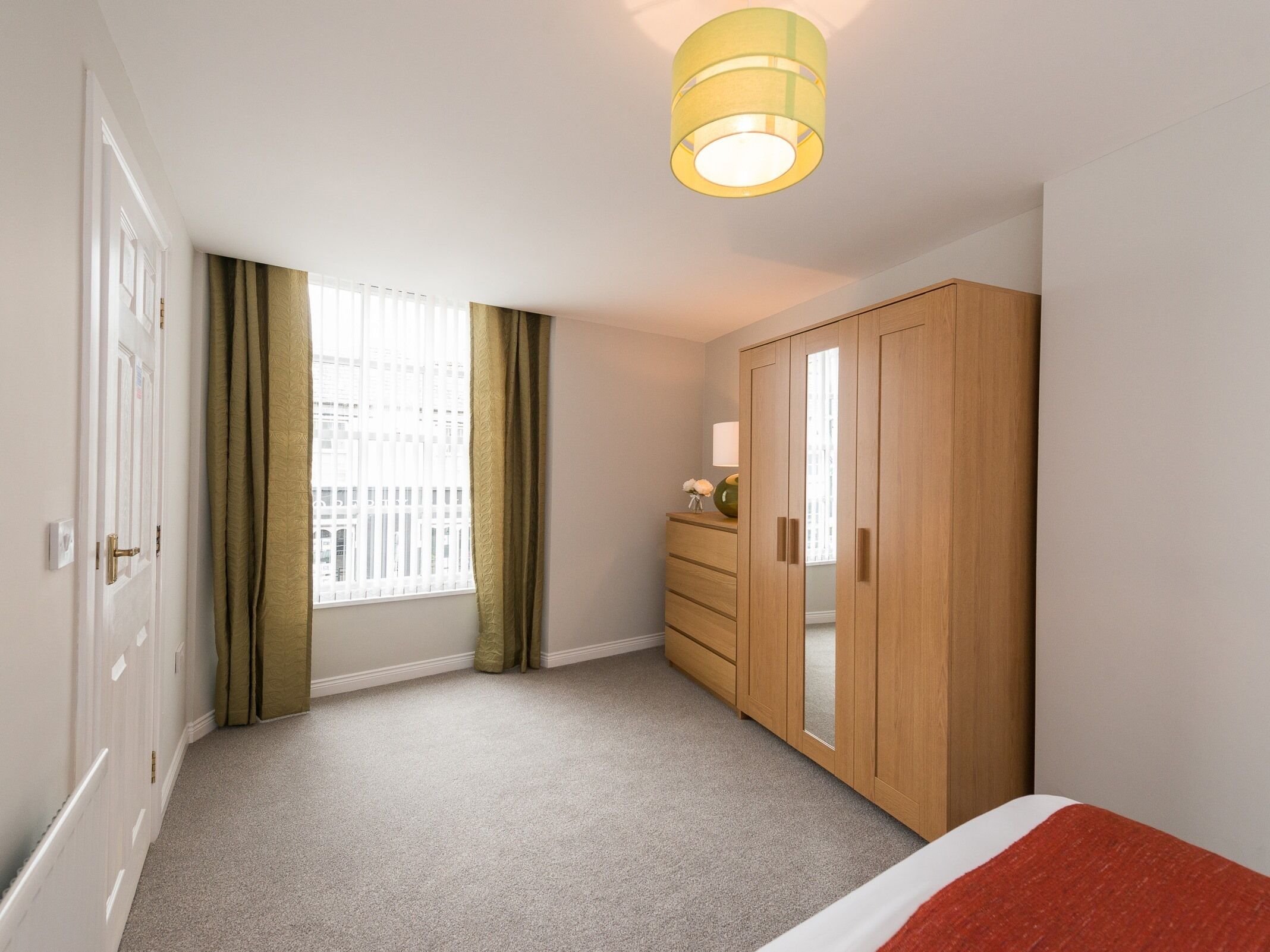 A bedroom with a large wardrobe, a bed with a red blanket, a window with curtains, and a ceiling light.