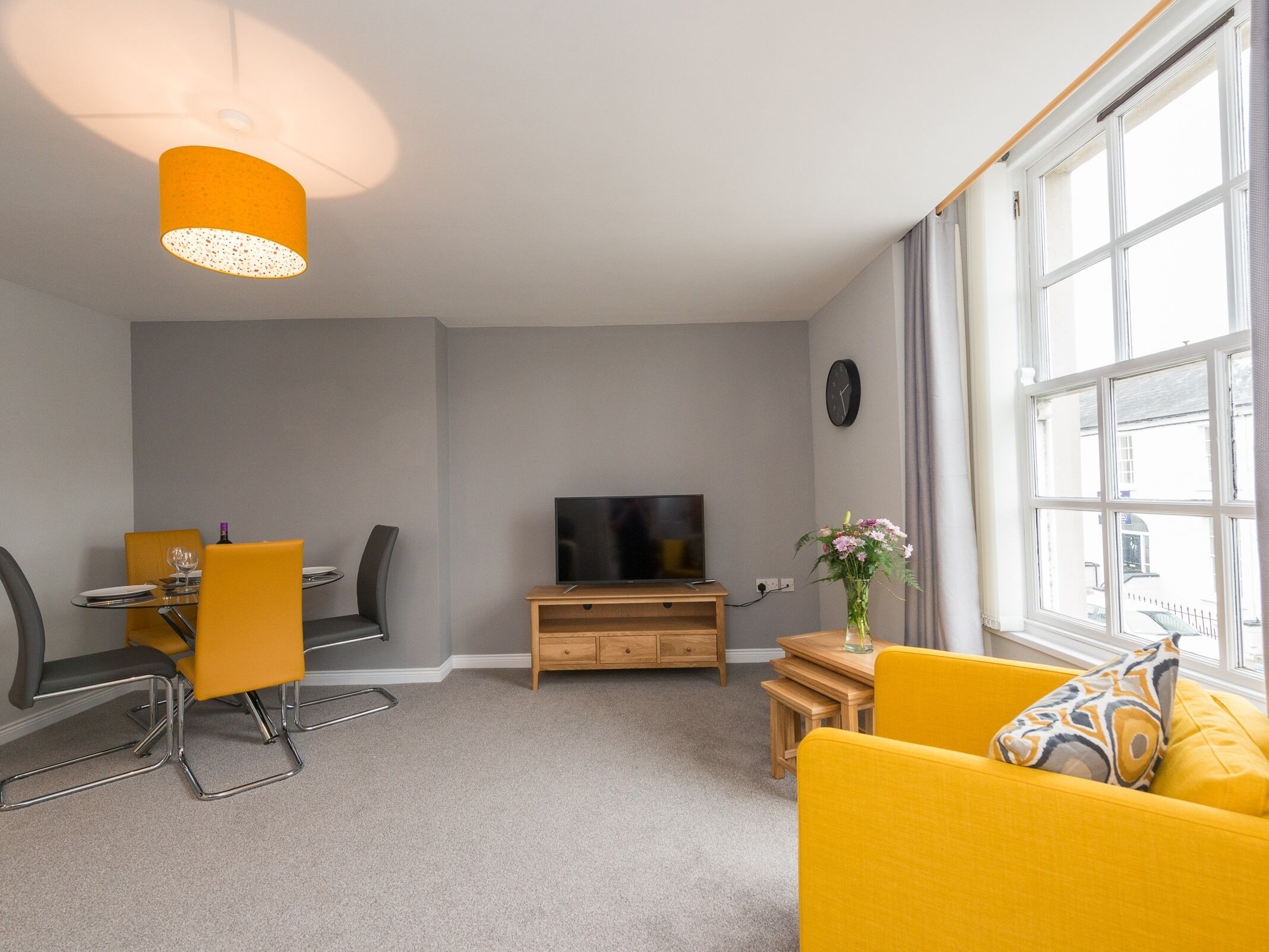 A modern living room with a dining area, a flat-screen TV, and a large window with a view of the street.