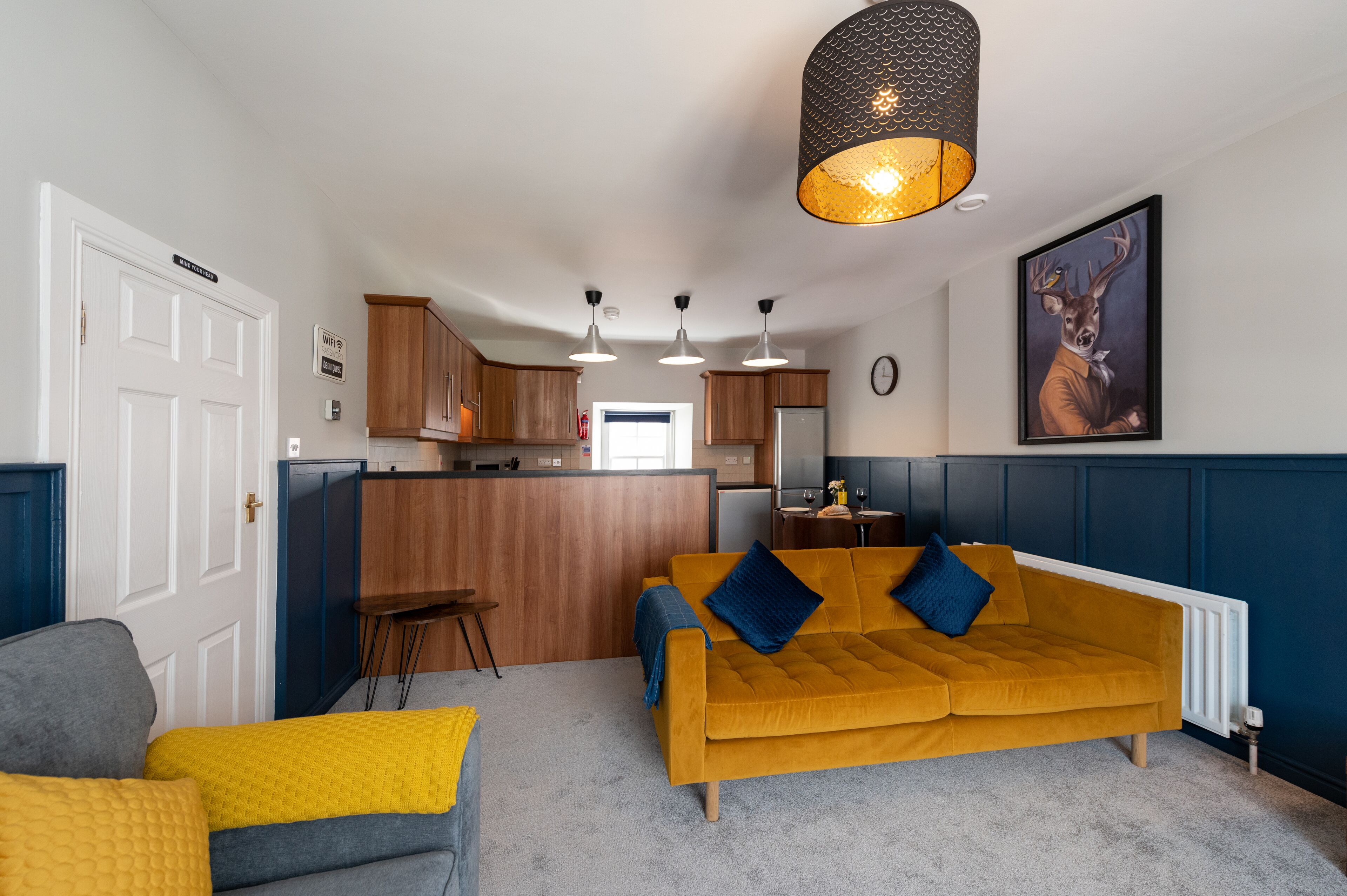 Una sala de estar moderna con un sofá color mostaza, un sillón gris y una manta amarilla. Se observa una zona de cocina con armarios de madera y un taburete de bar. Un cuadro grande de un ciervo cuelga en la pared.