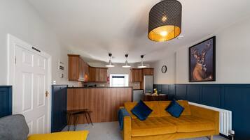Una sala de estar moderna con un sofá color mostaza, un sillón gris y una manta amarilla. Se observa una zona de cocina con armarios de madera y un taburete de bar. Un cuadro grande de un ciervo cuelga en la pared.