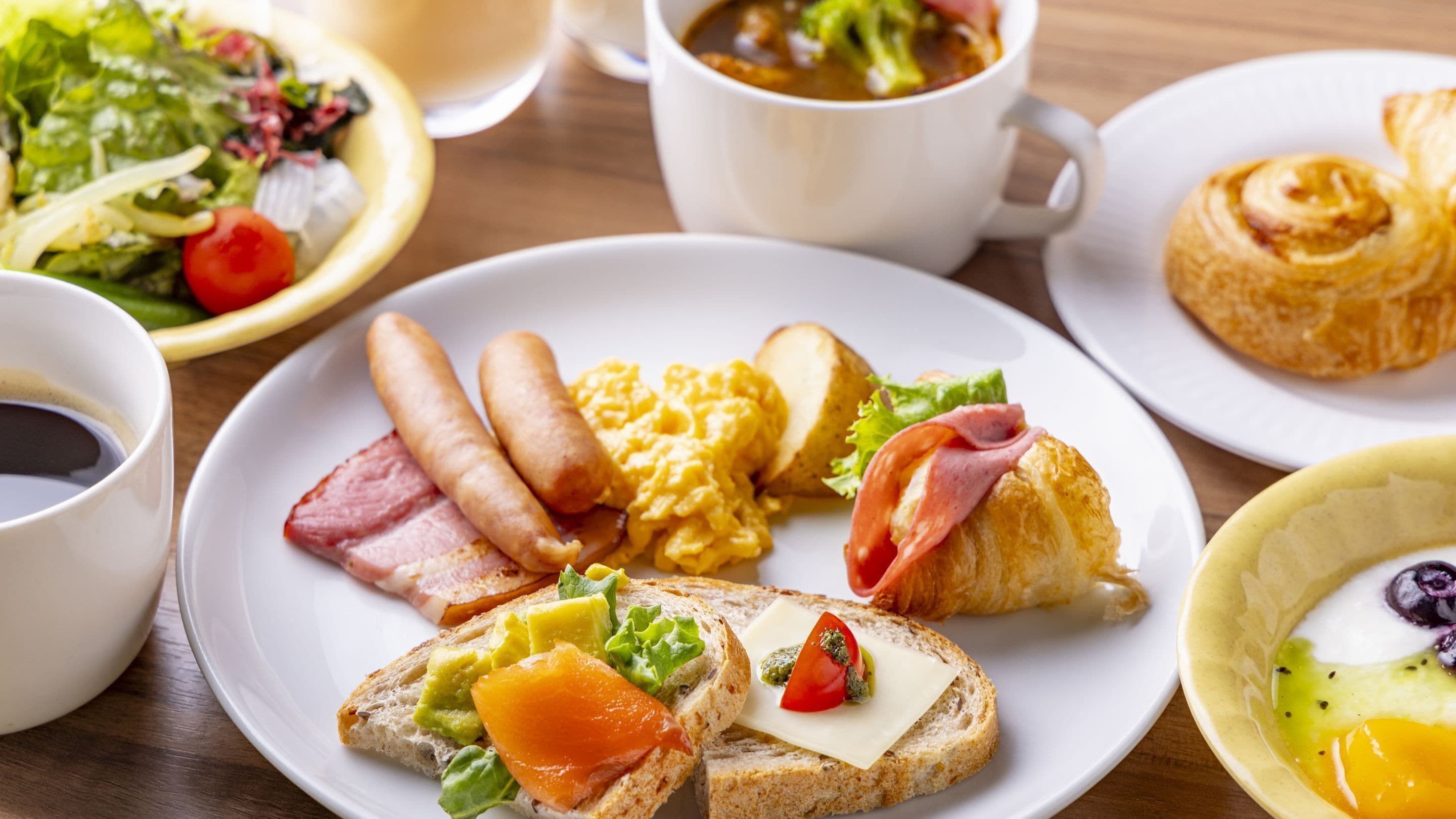 A breakfast setting with plates of eggs, bacon, sausages, toast with avocado and tomato, a croissant, and a cup of coffee.