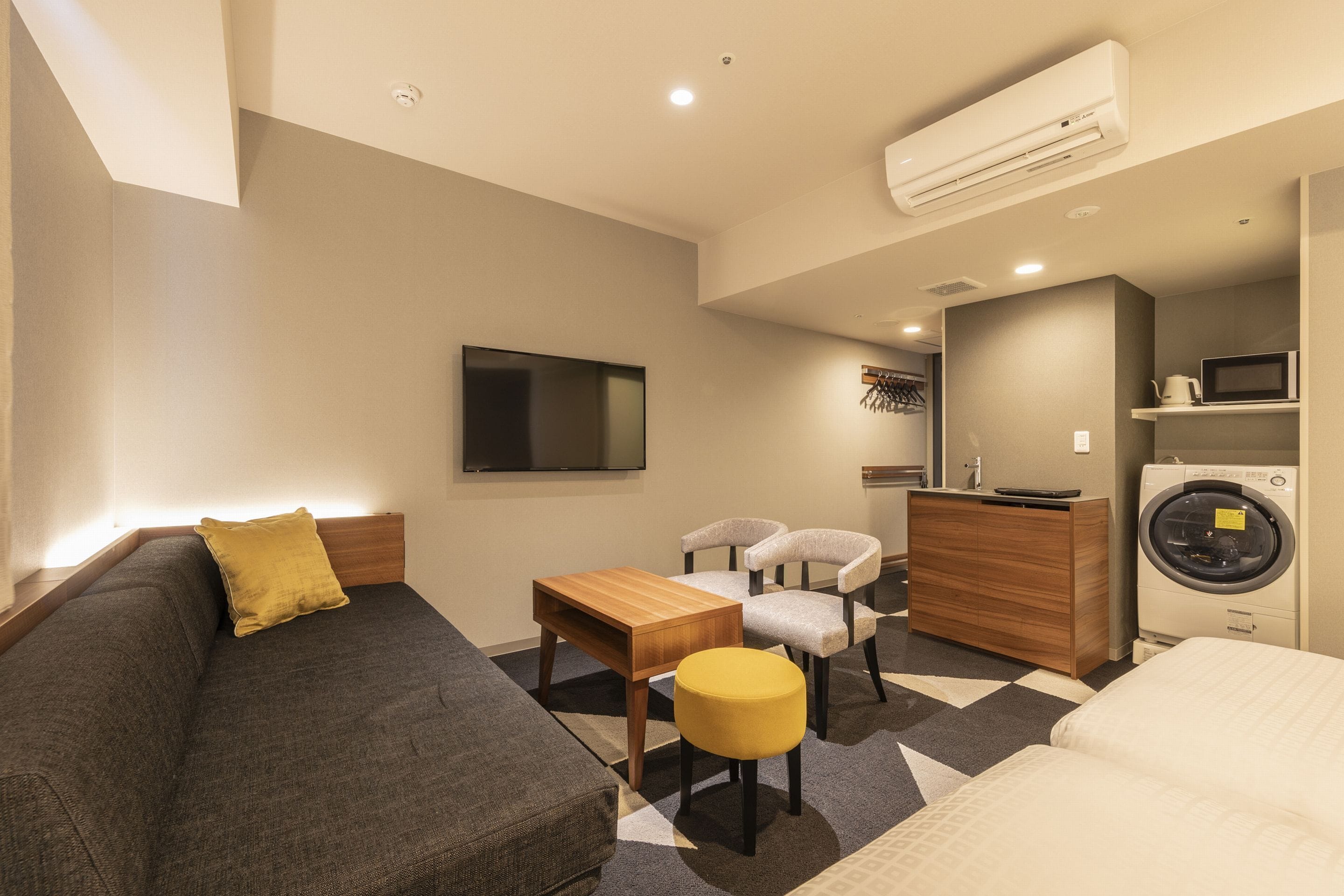 A modern hotel room with a sofa, a small table, two chairs, a TV, a washing machine, and a microwave.