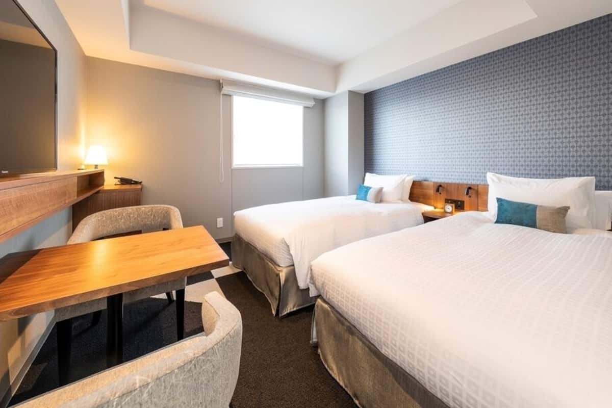 Une chambre d’hôtel avec deux lits, un bureau, une chaise, une télévision et une fenêtre.