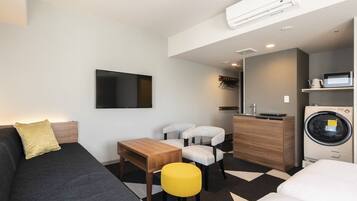 A modern hotel room with a sofa, a small table, two chairs, a bed, a washing machine, a TV, and a kitchenette.