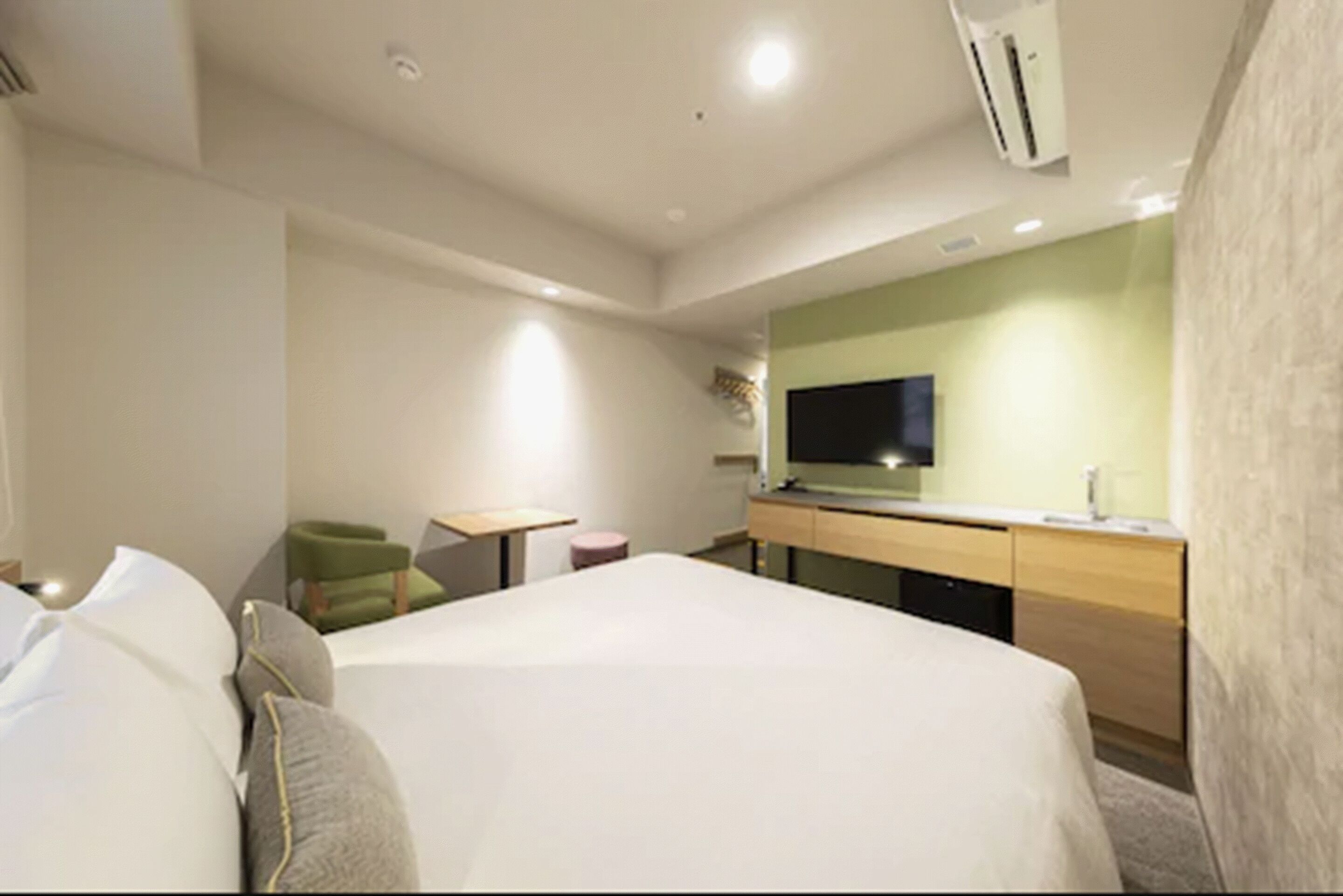 A modern hotel room with a large bed, a small desk, a chair, a television, and a minibar.