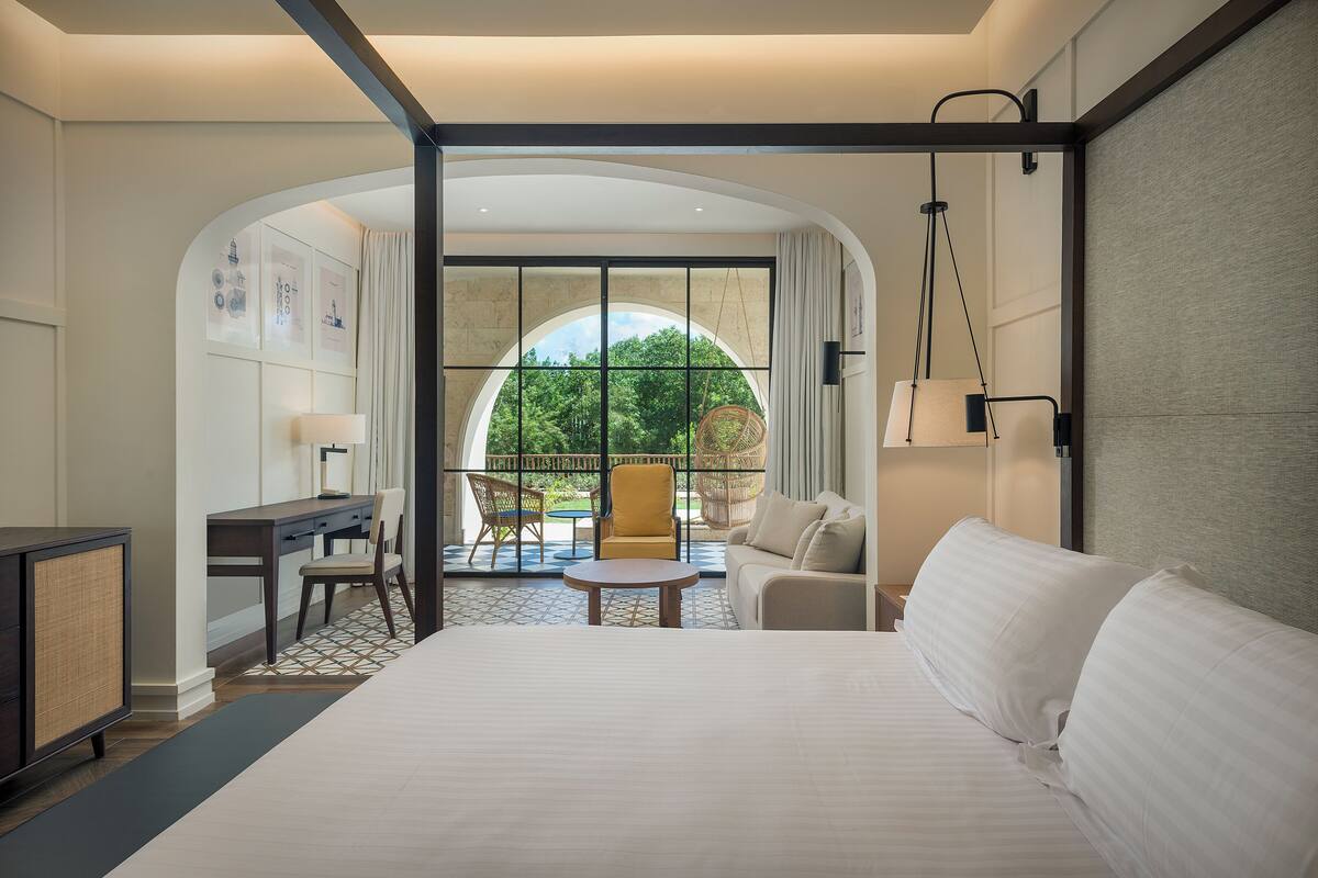 Une chambre d’hôtel avec un grand lit, un bureau avec une chaise, une petite table et un canapé. Une grande fenêtre offre une vue sur la verdure environnante.