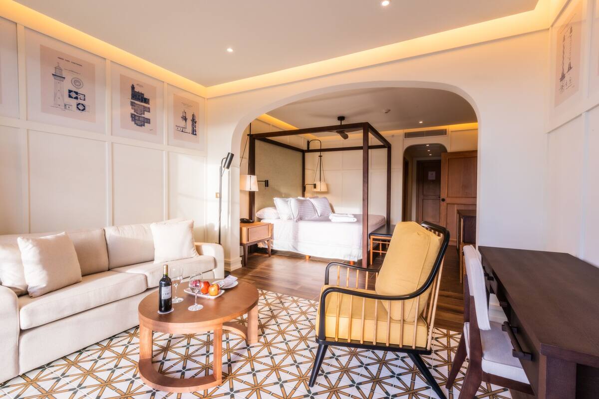 Une chambre d’hôtel comprenant un canapé, un fauteuil, une table à manger, un lit et une petite table sur laquelle se trouvent une bouteille et des verres.