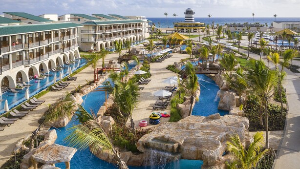 Resort com várias piscinas, incluindo uma grande piscina central com cascata, e área de praia com guarda-sóis.