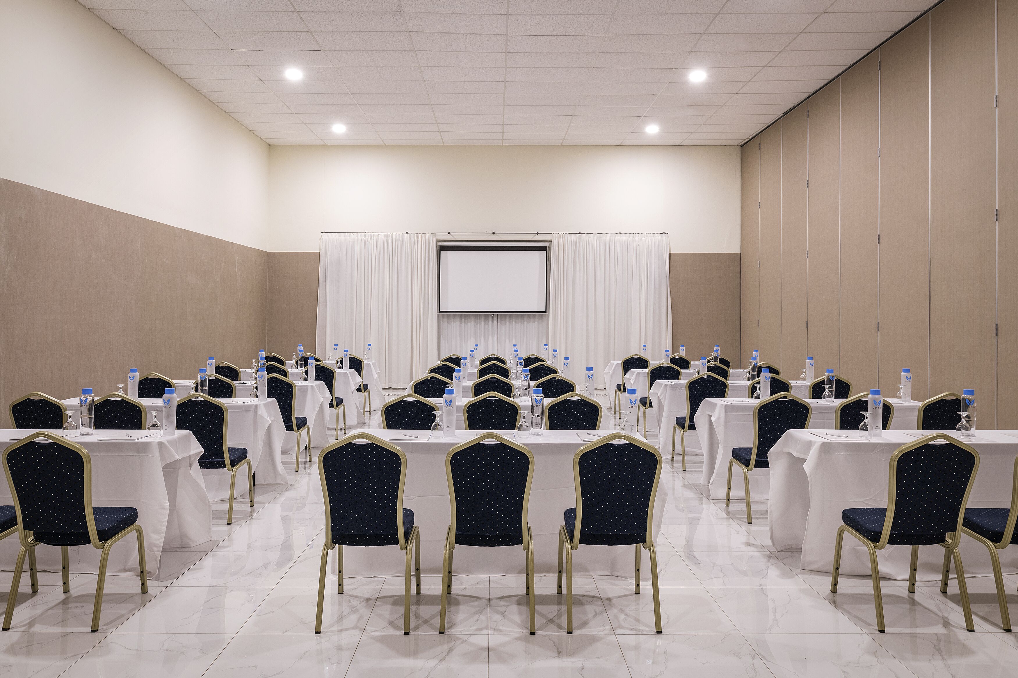 A conference room with rows of tables and chairs, a projector screen, and a white curtain backdrop.