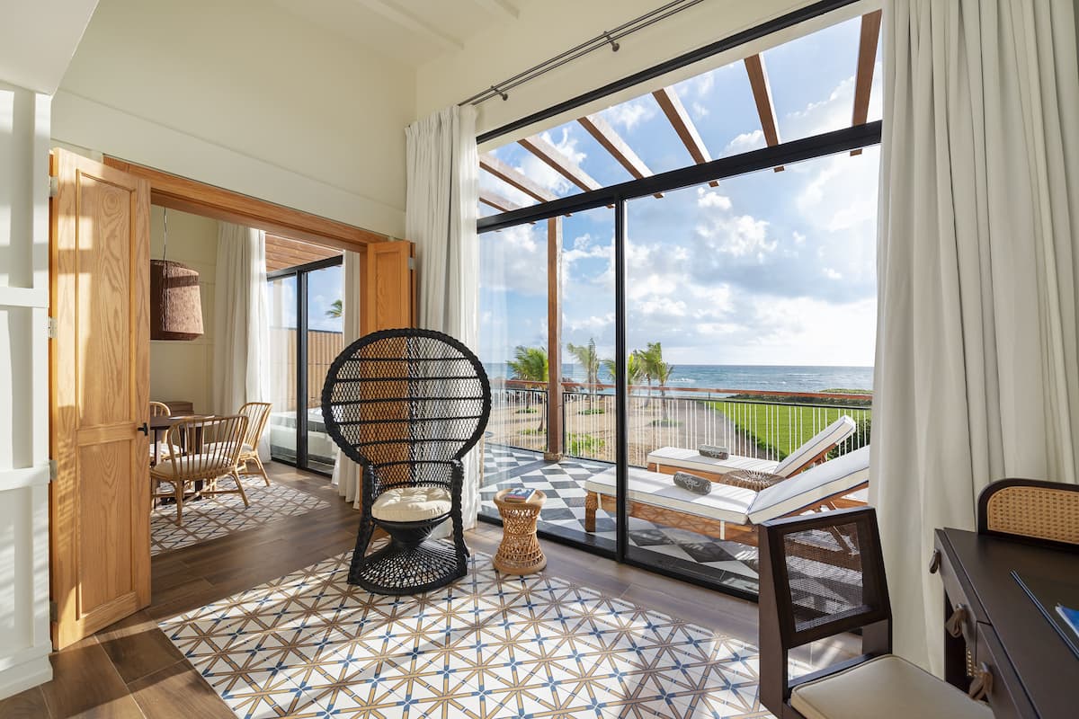 Une pièce dotée d’une grande fenêtre donnant sur une plage, d’un fauteuil en osier, d’une petite table et d’une vue sur l’océan.