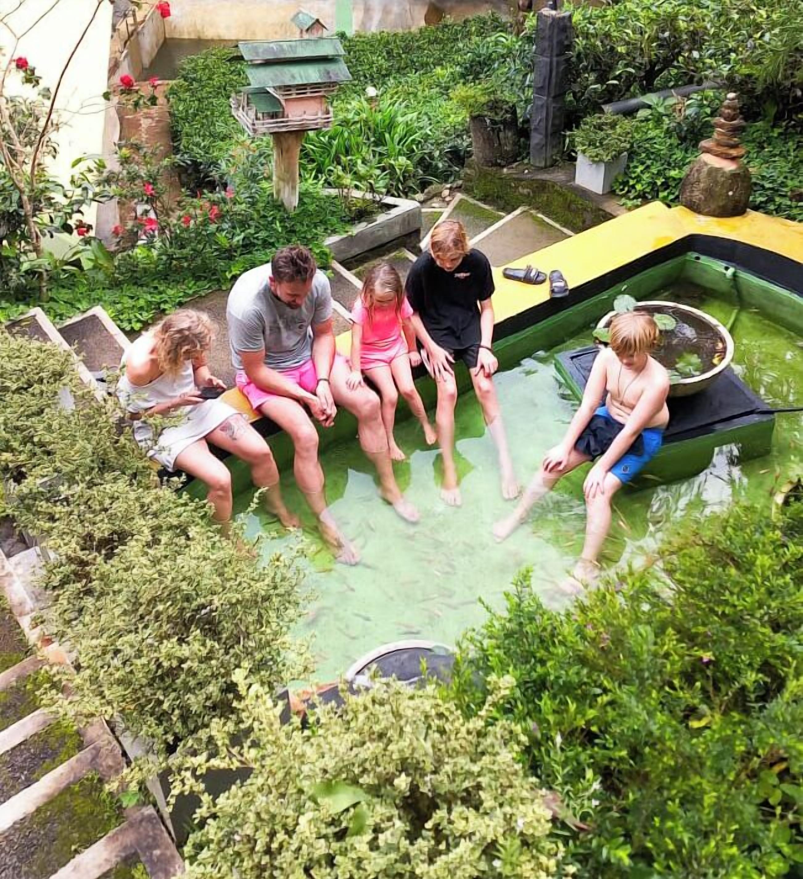 Un groupe de personnes est assis sur un bateau dans un jardin où l’on peut voir différentes plantes et un petit étang.