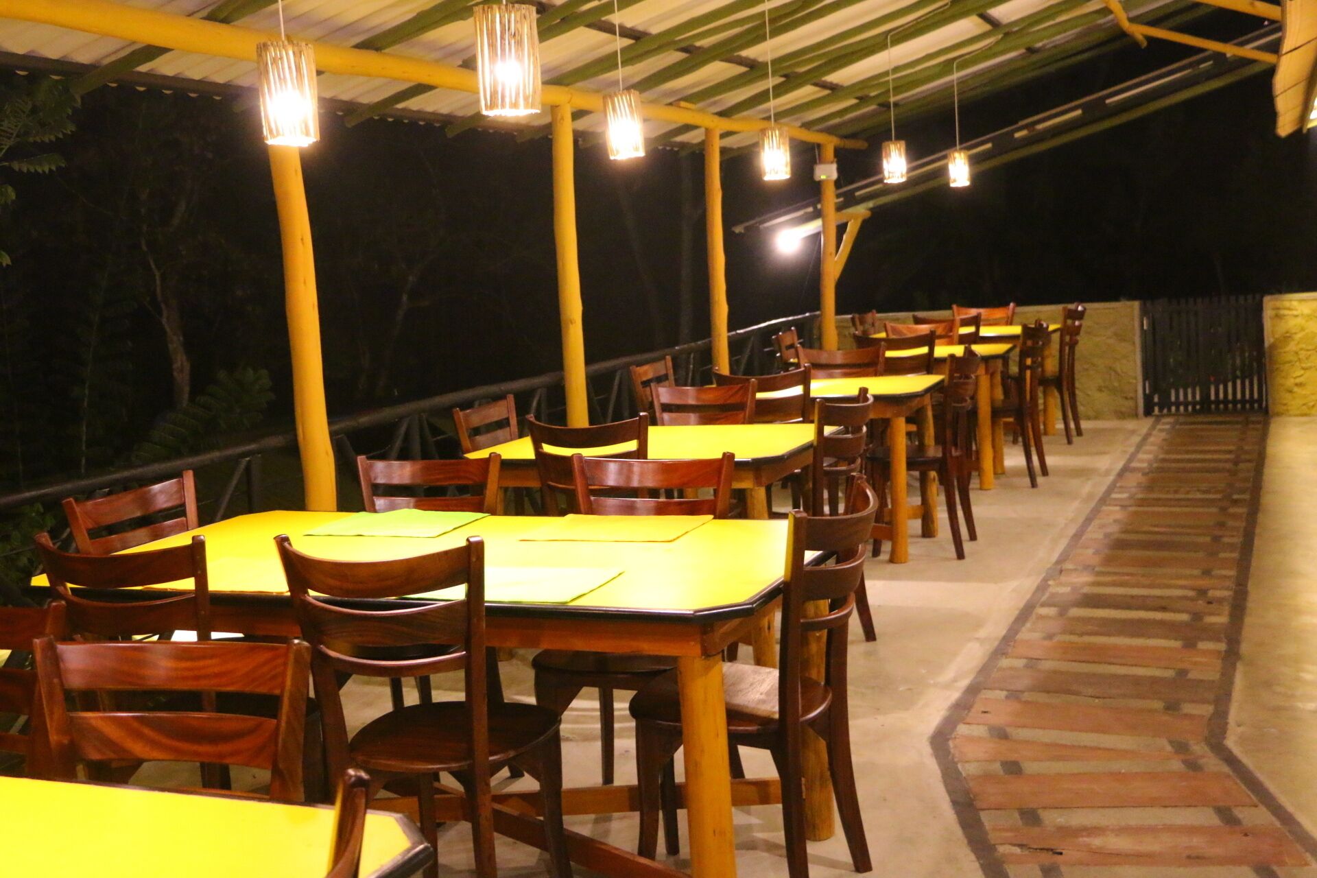 Un espacio para comer al aire libre con mesas y sillas de madera, bajo una estructura cubierta con luces colgantes.
