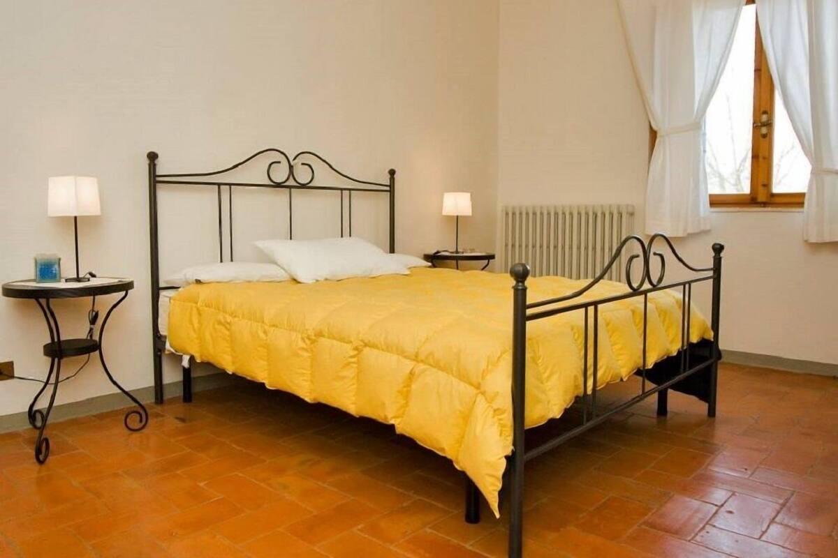 Una cama individual con estructura metálica, dos mesitas de noche con lámparas, un radiador y una ventana con cortinas.