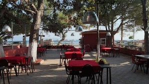An outdoor dining area with red tables and chairs, surrounded by trees and overlooking a body of water. - Deniz Kizi Otel (Armutlu)