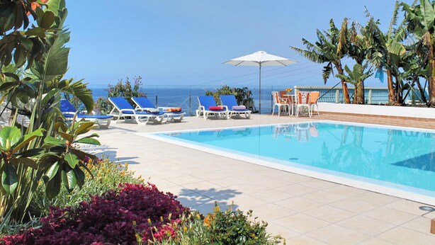 Zona junto a la piscina con sillones de descanso y una sombrilla, con vista a un cielo azul y al océano.