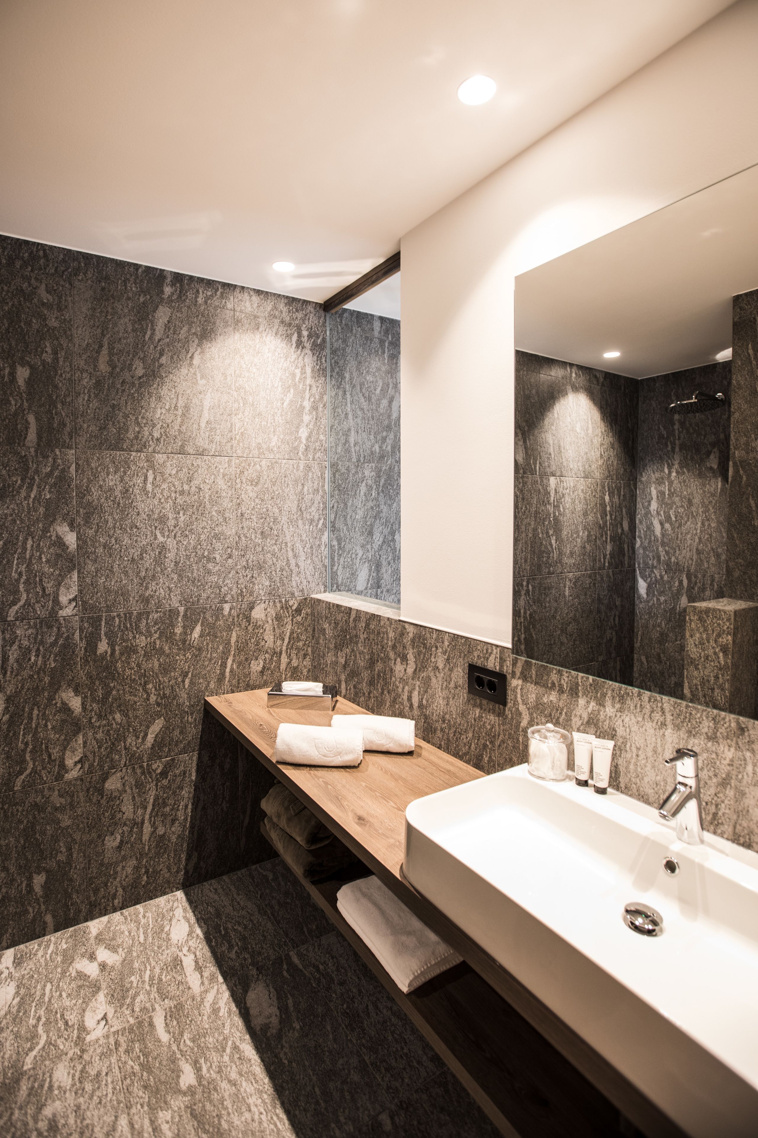 Une salle de bain moderne avec un mur en marbre, un meuble-lavabo en bois, un grand miroir et un lavabo blanc.