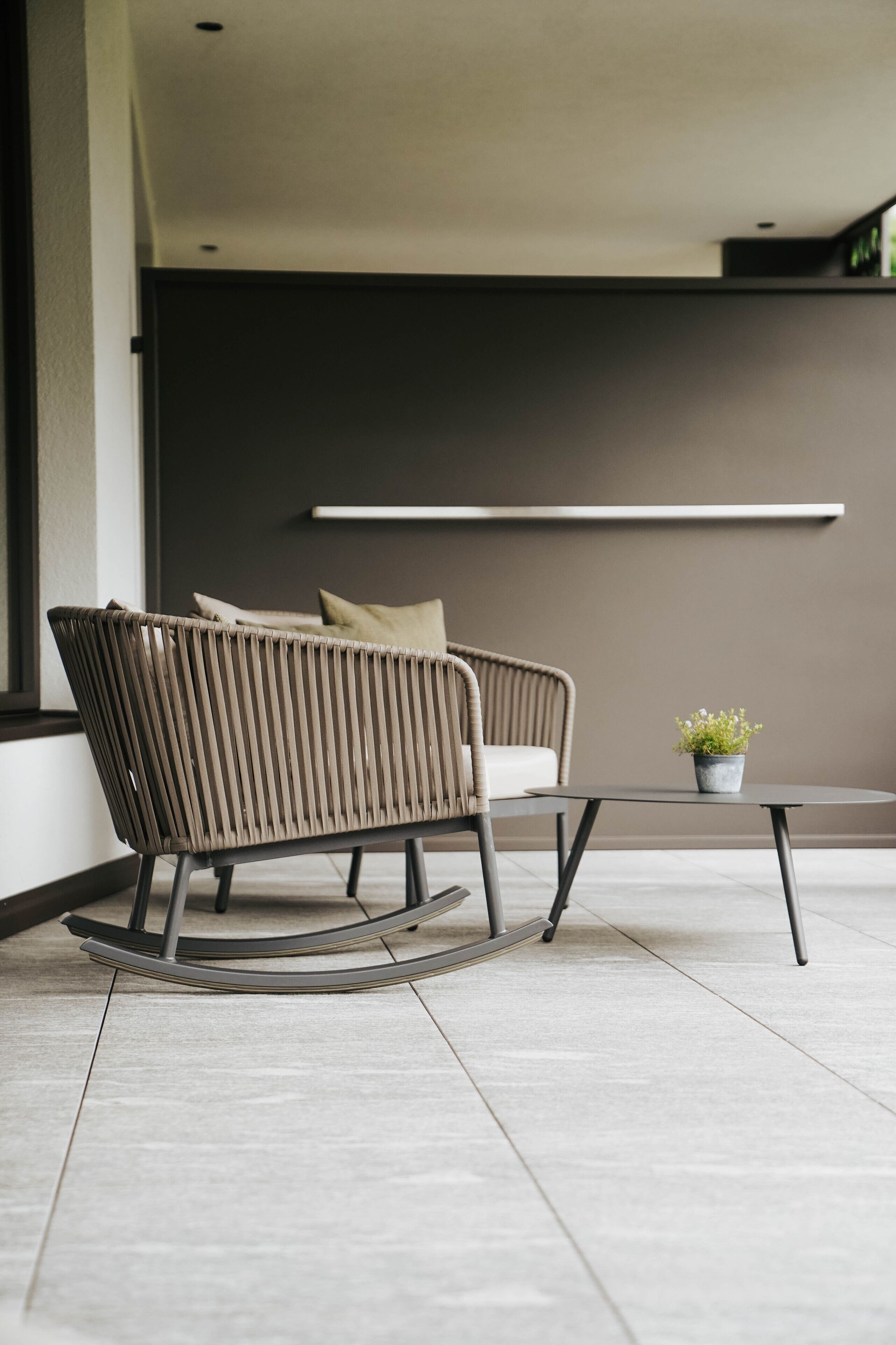 A modern outdoor seating area with a wicker chair and a small table, a potted plant, and a minimalist wall design.